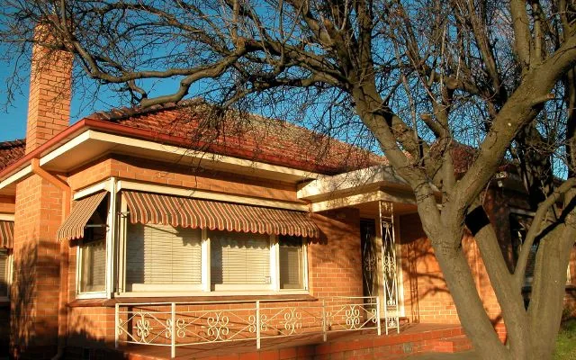 Red-brick residence with a striped window awning beneath mature trees.