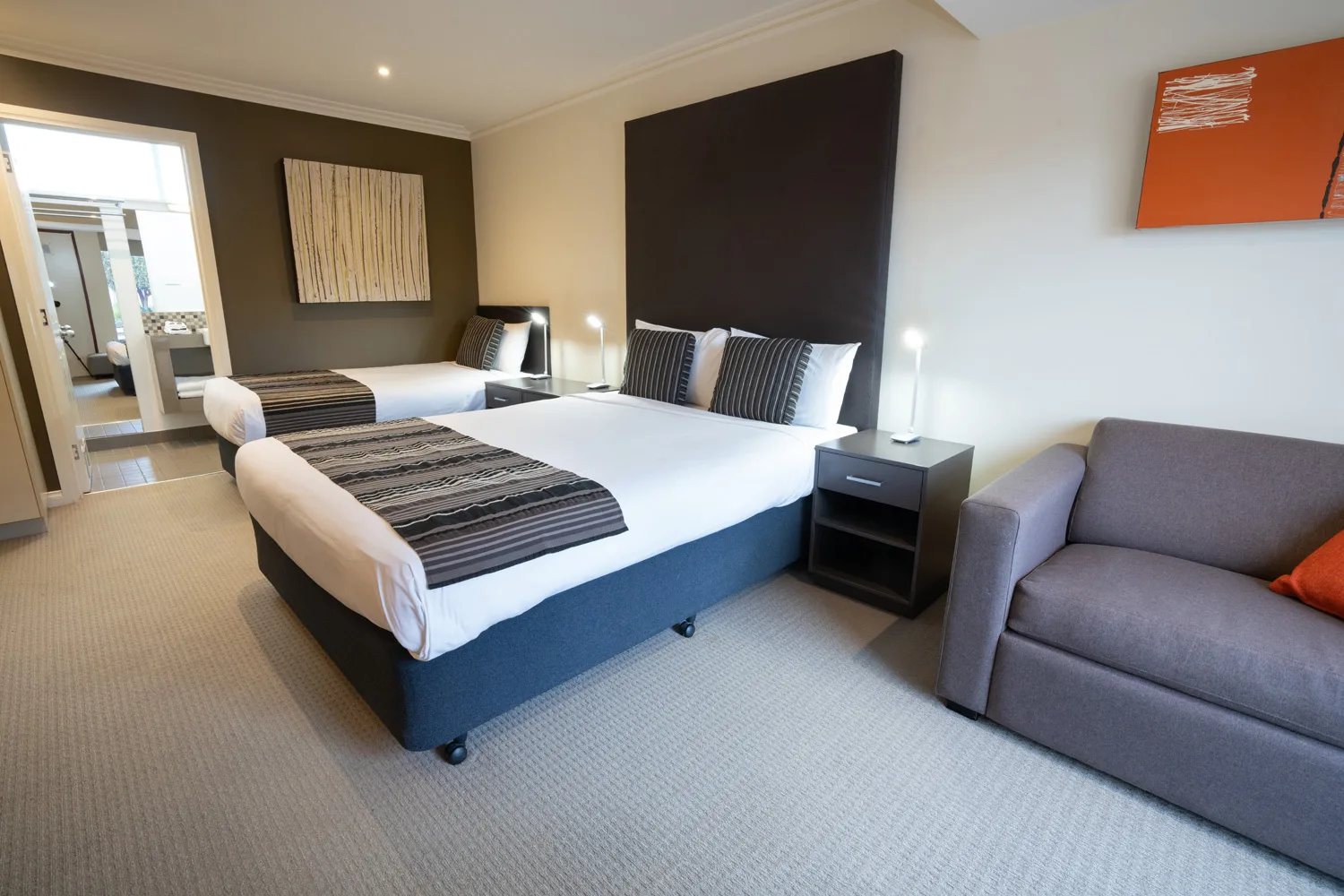 A motel room with two beds, a grey sofa and an ensuite bathroom.